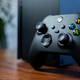 An Xbox Series X controller rests against an Xbox Series X on a table.